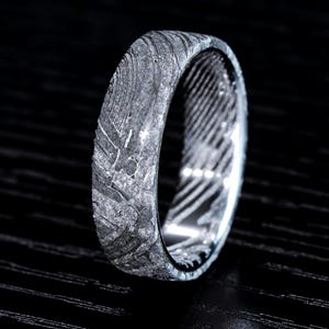 May include: A close-up of a silver ring with a unique, textured pattern. The ring's surface displays a marbled effect with shades of gray and silver, creating a striking visual appeal. The ring is set against a dark background.