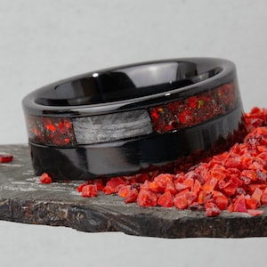 May include: A black ring with a red and gray inlay. The ring is sitting on a bed of red crushed glass.