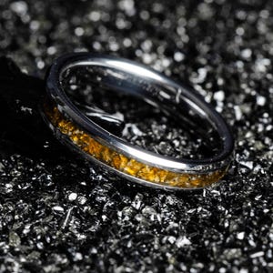 May include: A silver ring with a band of amber-colored stones. The ring is set against a dark, textured background, highlighting the contrast between the shiny metal and the rough surface.