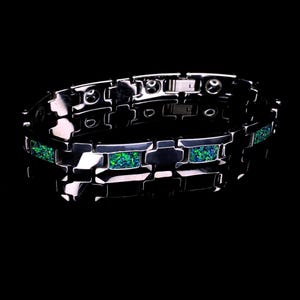 Puede incluir: Una pulsera de eslabones en tono plateado pulido con incrustaciones rectangulares de piedra triturada iridiscente en tonos verde y azul. La pulsera está formada por segmentos metálicos entrelazados con un cierre visible en la banda interior.