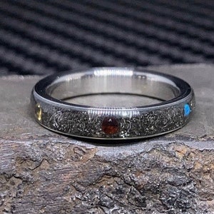 Puede incluir: Un anillo de plata con una banda oscura y texturizada, con pequeñas piedras preciosas de colores incrustadas. El anillo presenta una piedra roja, azul y amarilla, y está sobre un fondo texturizado. Es una joya.