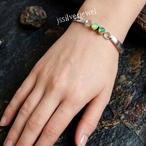 Peut inclure: Un bracelet manchette en argent orné de deux pierres d'opale rondes et irisées serties au centre. La bande métallique présente une finition lisse et polie avec des maillons circulaires reliant les pierres au corps. Le texte jssilverjewel est visible.