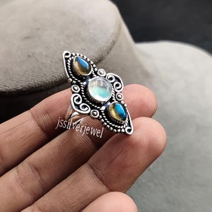 Può includere: Un anello d'argento con tre pietre preziose. La pietra centrale, rotonda, è iridescente, affiancata da due pietre a forma di goccia. L'anello presenta un ornato design a filigrana con il testo "jssilverjewel".