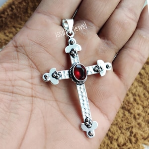 Puede incluir: Pendiente de cruz de plata con una piedra preciosa roja en el centro. La cruz tiene una superficie texturizada y detalles ornamentados.