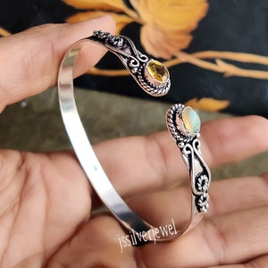 May include: Silver bangle bracelet with an ornate design featuring two gemstones. One gemstone is a yellow citrine and the other is a white opal.