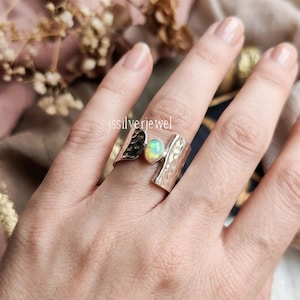 May include: A wide silver ring featuring a central oval-shaped iridescent opal stone. The band has a textured, hammered finish on both sides of the stone. The text jssilverjewel is visible on the metal surface of the ring band.