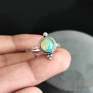 Puede incluir: Un anillo de plata con una piedra preciosa de ópalo redonda e iridiscente engastada en un engaste de bisel. El anillo tiene un diseño decorativo con pequeñas cuentas alrededor de la piedra preciosa.