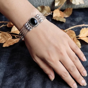 May include: A silver cuff bracelet featuring a central oval black stone set in a decorative metal frame. The band consists of multiple thin silver wires that converge at the sides. The text jssilverjewel is visible above the bracelet.