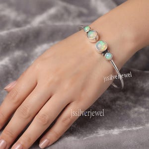 Peut inclure: Un bracelet jonc en argent orné de quatre pierres d'opale rondes et irisées serties dans des chatons en métal lisse. Le bracelet est composé d'une fine tige en argent poli qui épouse le poignet. Le texte jssilverjewel est visible sur l'image.