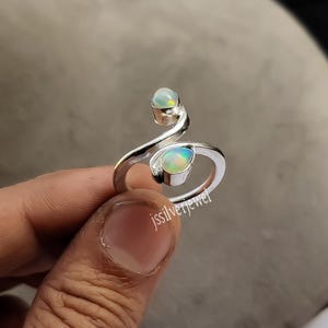 May include: A silver ring featuring two opal gemstones. The ring has a distinctive, swirling design with one opal at the top and another at the bottom. The opals display a mix of colours, including green, blue, and yellow.