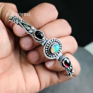 May include: A silver bangle bracelet with three gemstones: a black stone, a white opal, and a red garnet. The bracelet is decorated with intricate silver detailing.