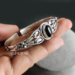 May include: Silver bangle bracelet with an ornate design and a black oval gemstone. The bracelet is open and adjustable.