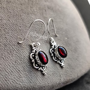 May include: Silver earrings featuring oval, dark red gemstones. The earrings have an ornate design around the gemstones and a curved hook. The text "jssilverjewel" is visible on the left earring. A pair of elegant earrings.