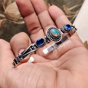 May include: Silver bangle bracelet featuring three oval-shaped gemstones. The central stone is a white opal, while the two side stones are blue. The bracelet has a delicate, ornate design.