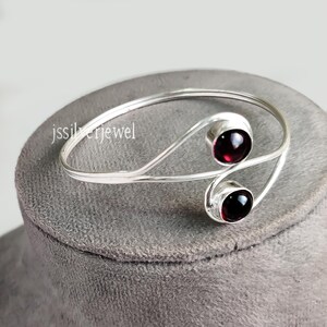 以下が含まれることがあります： 2つの赤い宝石のアクセントが付いたシルバーのバングルブレスレット。ブレスレットは開いており、湾曲したデザインになっています。