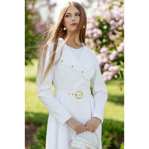 May include: A white long-sleeved dress with a belted waist and a gold buckle. The dress has a collar and a button closure. The model is wearing the dress with a white handbag.