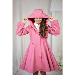 May include: A pink hooded raincoat with red floral embroidery. The coat features a flared skirt and a button-up front. The hood is up, and the lining is red. The sleeves are long, and the coat is worn over a white collared shirt.