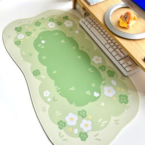May include: A light green mousepad with a whimsical garden design. The pad features a central green area surrounded by a lighter green border with white flowers and green clovers. The design is reminiscent of a meadow.