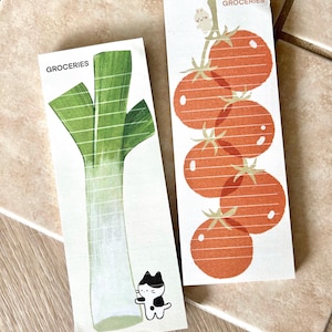 May include: Two rectangular notepads with illustrations of groceries. One features a leek, the other a cluster of tomatoes. Both have the word "GROCERIES" at the top. A small black and white cat is on the leek notepad.