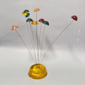 May include: A decorative sculpture featuring a yellow, translucent, dome-shaped base with multiple thin, dark metal wires extending upward. Each wire is topped with a small, colorful, translucent, dome-shaped cap in shades of red, orange, and blue.