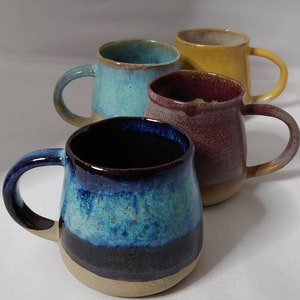 May include: Four ceramic mugs with different coloured glazes. The mugs are all the same size and shape, with a rounded body and a handle. The glazes are a light blue, a dark purple, a yellow, and a blue and black.