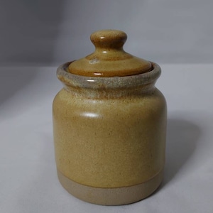 Puede incluir: Un pequeño tarro redondo de cerámica con tapa. El tarro es de color marrón claro con un esmalte marrón más oscuro. La tapa es de color marrón más oscuro con un esmalte marrón más claro.