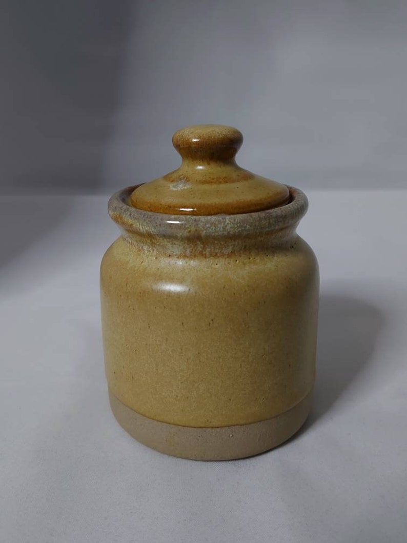 Puede incluir: Un peque&ntilde;o tarro redondo de cer&aacute;mica con tapa. El tarro es de color marr&oacute;n claro con un esmalte marr&oacute;n m&aacute;s oscuro. La tapa es de color marr&oacute;n m&aacute;s oscuro con un esmalte marr&oacute;n m&aacute;s claro.