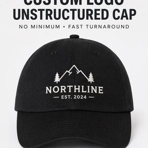 Puede incluir: Gorra negra sin estructura con el texto "CUSTOM LOGO UNSTRUCTURED CAP" en la parte superior. La gorra presenta un logotipo blanco de montaña y árboles con la palabra "NORTHLINE" y "EST. 2024" debajo. La gorra está hecha de un material suave.