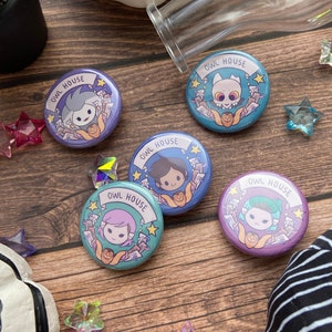May include: Four round purple and blue buttons with a white border and gold stars. Each button features a different character from the animated series "The Owl House" with the text "Owl House" on a banner.