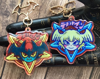 May include: Two colorful keychains featuring anime characters with Japanese text. The keychains are made of clear acrylic and have a starburst design. The first keychain features a red and blue demon with bat wings. The second keychain features a blonde girl with angel wings.