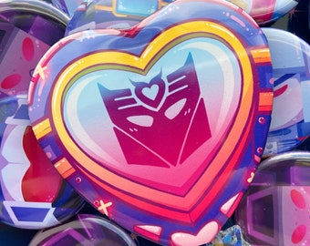 May include: A heart-shaped button with a rainbow border and a pink heart in the center. The heart contains a stylized purple and pink robot face with a smaller pink heart in the center of the face.