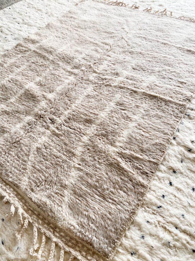 Pu&ograve; includere: Un tappeto color crema con una texture soffice e frange. Il tappeto &egrave; fatto di lana e ha un aspetto naturale e rustico.
