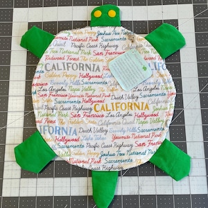 Puede incluir: Un juguete de suelo hecho a mano con forma de tortuga con aletas, cabeza y cola verdes. El caparazón de la tortuga es blanco y está cubierto de texto colorido con monumentos de California. El juguete tiene dos ojos de botón amarillos y una pequeña etiqueta.