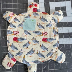 May include: A turtle-shaped kitchen floor mat with a cream background and a pattern of gray pigeons, red houses, and red and white striped accents. The mat has a red stitched border and the text "Floor Turtle".