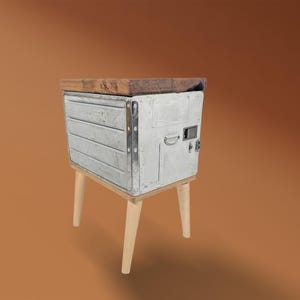 Op de afbeelding: Een kleine bijzettafel met een lichtgrijze metalen kast en een donkerhouten blad. De tafel heeft vier schuine, lichtgeleurde houten poten. Het ontwerp is een mix van rustieke en moderne elementen. Afmetingen: ongeveer 40 cm hoog.