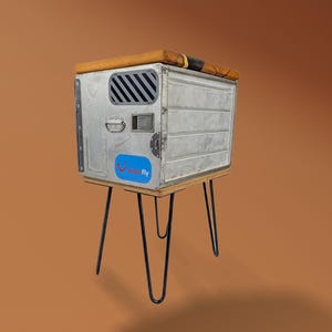 Op de afbeelding: Een unieke bijzettafel gemaakt van een hergebruikte metalen container. De tafel heeft een houten blad, zwarte hairpinpoten en een blauw-witte sticker. De container heeft een ventilatie en een handvat.