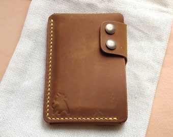 May include: A brown leather wallet with a flap closure and two silver snaps. The wallet is sitting on a white fabric surface.