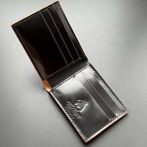Könnte beinhalten: Ein schwarzes Bifold-Portemonnaie mit glänzender Oberfläche und hellbraunen Kanten. Das Innere verfügt über mehrere vertikale Kartenfächer und ein eingeprägtes Logo mit der Aufschrift "Toro Negro crafted by hand" sowie sichtbare Nähte.