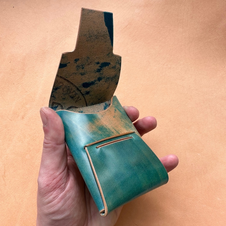 Op de afbeelding: Een teal leren portemonnee met een klep die opengaat om een ​​tan leren interieur te onthullen. De portemonnee heeft een klein vakje aan de voorkant.