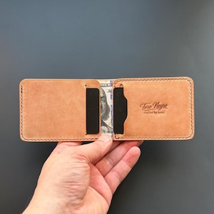 May include: A tan leather bifold wallet featuring black stitching and two black card slots. The interior displays a partial United States one hundred dollar bill. The right side is embossed with the text Toro Negro crafted by hand.