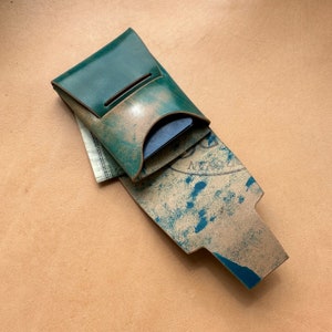Op de afbeelding: Een teal leren portemonnee met een klep en een versleten, tan leren interieur. De portemonnee is open en toont een blauwe kaart en een gevouwen biljet erin.