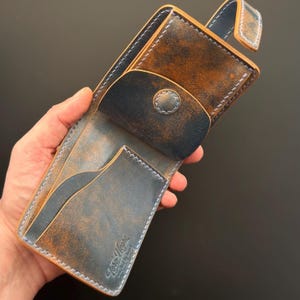 Può includere: Un portafoglio in pelle pieghevole con una finitura usurata blu e marrone e cuciture chiare. Il portafoglio include una chiusura a bottone automatico, diversi scomparti per carte e il testo in rilievo Toro Negro crafted by hand.