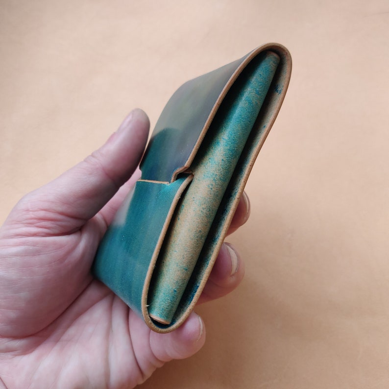 Op de afbeelding: Een teal leren portemonnee gevouwen en dicht, vastgehouden in een hand. De portemonnee heeft een lichtbruine leren voering.