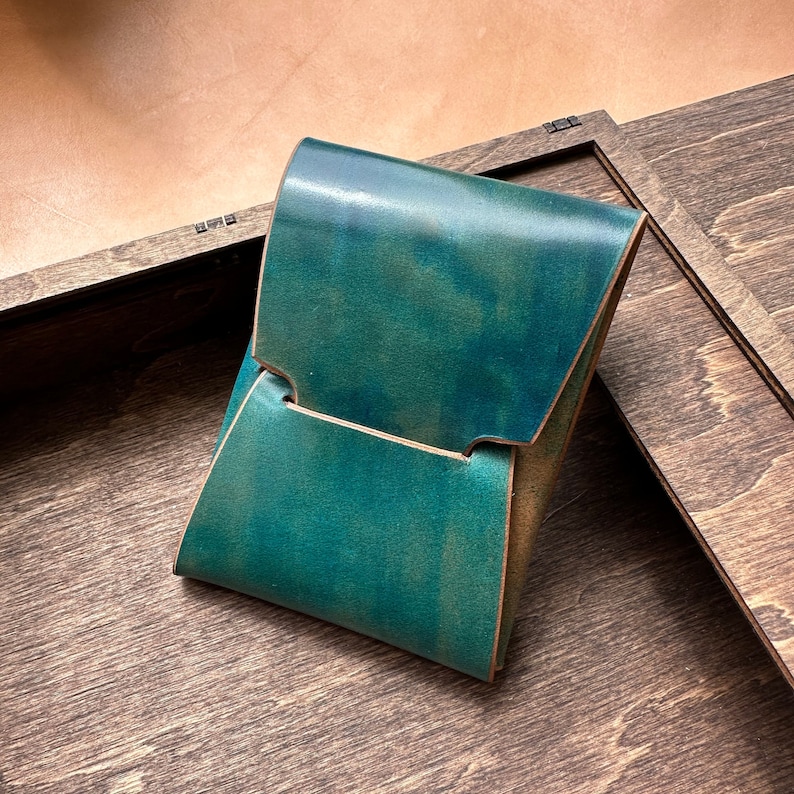Op de afbeelding: Een teal leren portemonnee met een klep en &eacute;&eacute;n vak. De portemonnee ligt op een houten oppervlak.