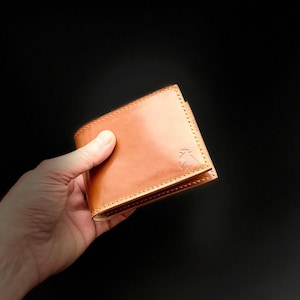 May include: A light brown leather wallet with a stitched border. The wallet is held in a hand against a black background. A small embossed design is visible on the front of the wallet.
