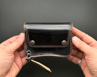 May include: A black leather wallet with a red stitch detail and two silver snaps. The wallet has a small flap with a cream-colored leather strap.