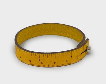 Op de afbeelding: Een gele leren armband met een liniaal erop gedrukt. De liniaal loopt van 1 tot 9 inch.
