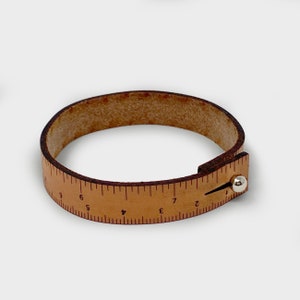 May include: A brown leather bracelet with a ruler printed on it. The ruler goes from 2 to 9 inches.