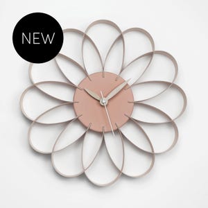 Peut inclure: Une horloge murale avec un centre circulaire rose poudré et douze boucles en forme de pétales s'étendant vers l'extérieur pour former une silhouette de fleur. Elle possède deux aiguilles rose poudré et une fine trotteuse argentée. Le texte NEW est présent.