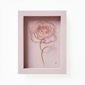Könnte beinhalten: Eine Rosenskulptur aus Kupferdraht in einem hellrosa Rahmen. Die Rose ist eine einzelne Blüte mit einem Stiel und Blättern.